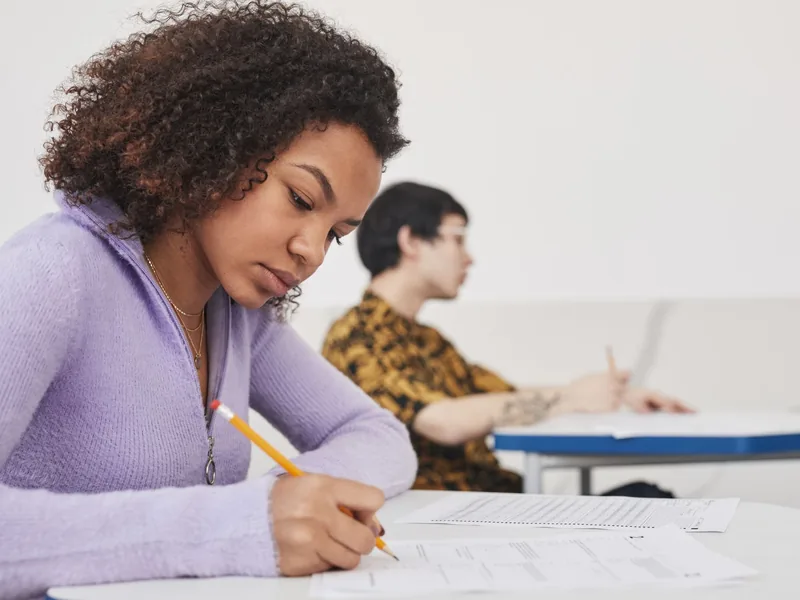 A student writing answers on paper at a desk in a classroom