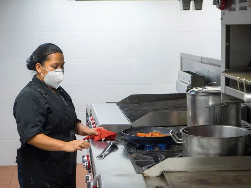 A cook in a face mask and black uniform working at the stove of a commercial kitchen