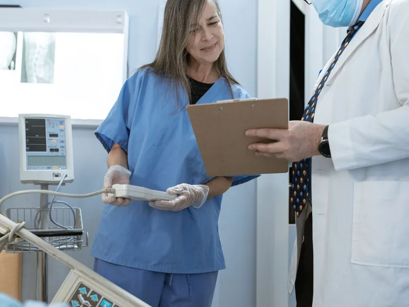 A nurse in blue scrubs writing on a clipboard beside a patient, with a doctor next to her