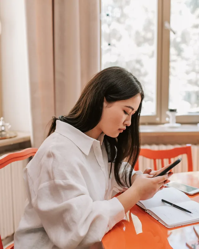A young woman at a window table, checking her phone while she writes notes in a notebook